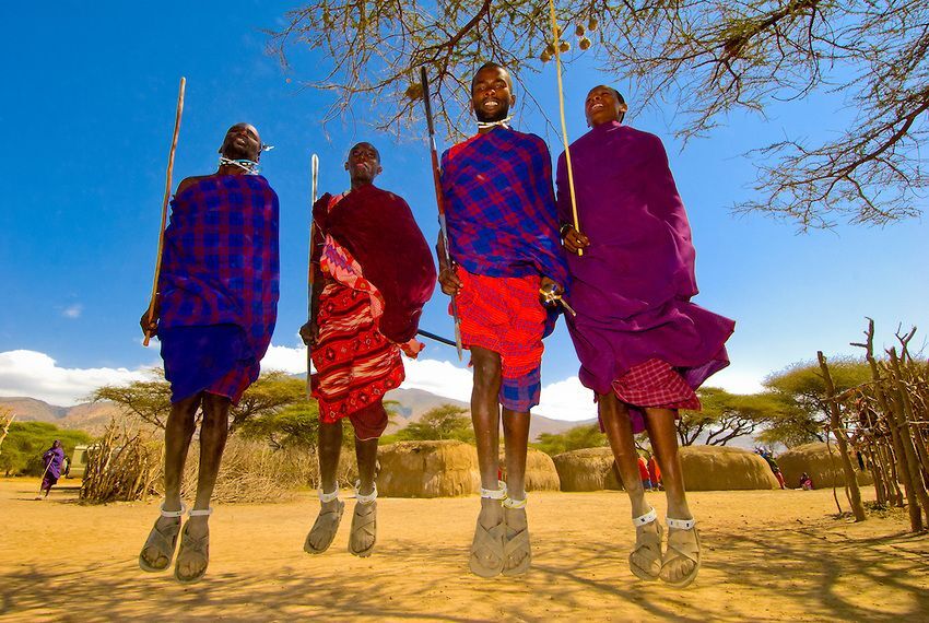Masai stam die de lucht in springt. Op de achtergrond zie je de typische Masai huisjes