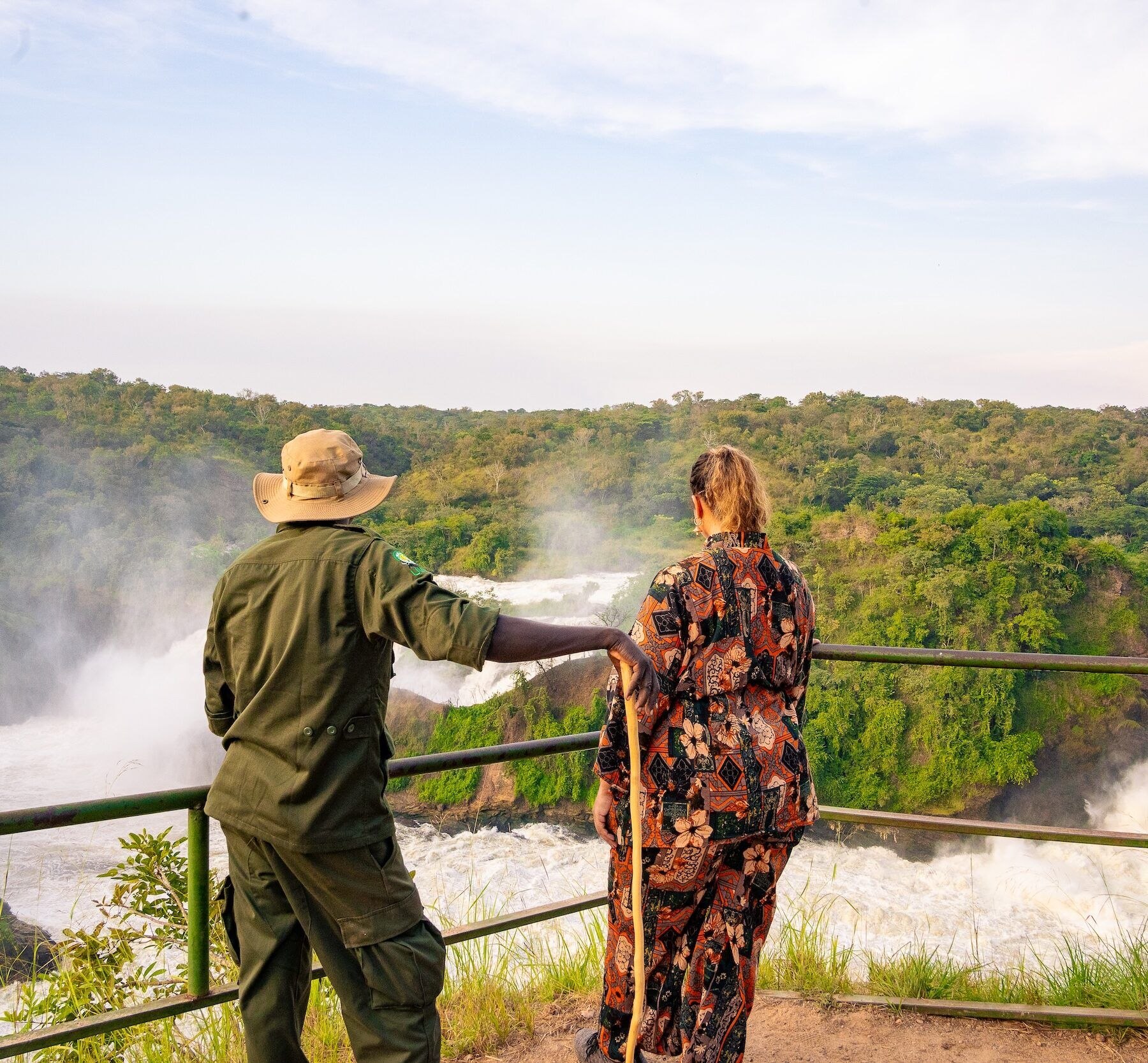 Een gids en een reiziger die de murchison falls bekijken vanaf een uitkijkpunt, met groene heuvels op de achtergrond.